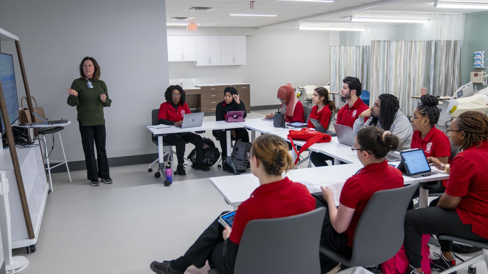 A group of nursing students attend lab class. Students sit in U shape facing a screen while professor lectures.