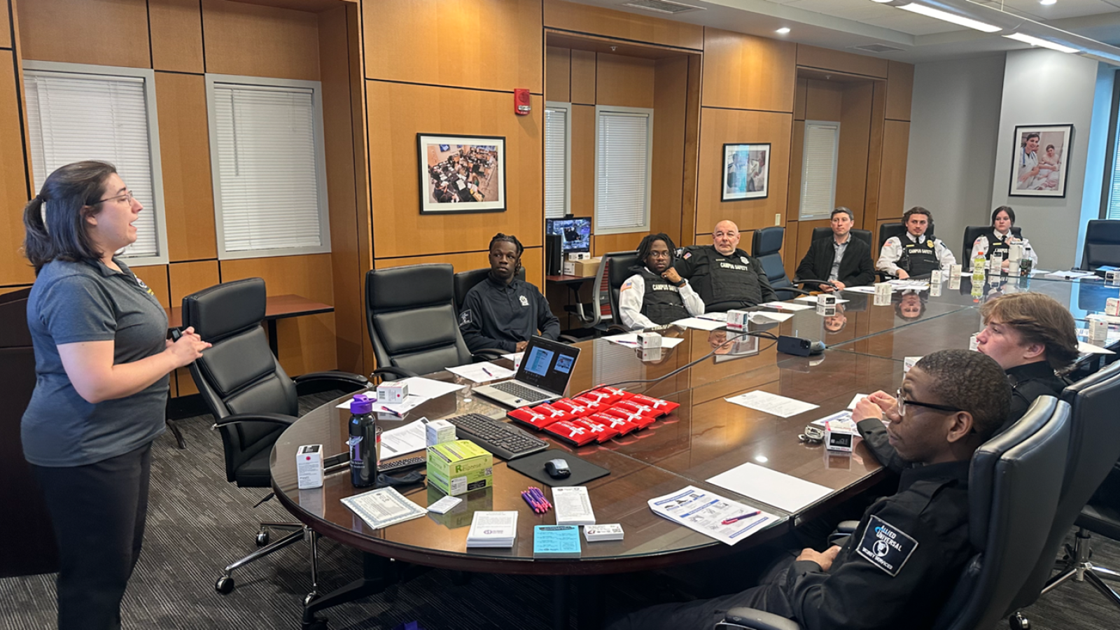 Group of police officers sit at a large conference table. A woman is standing at the head of the table speaking to the group.