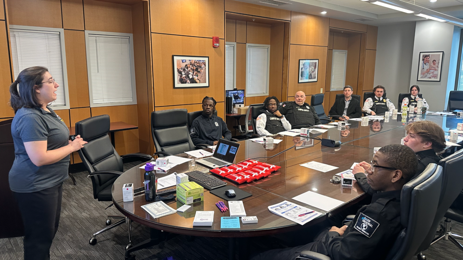 Group of police officers sit at a large conference table. A woman is standing at the head of the table speaking to the group. 