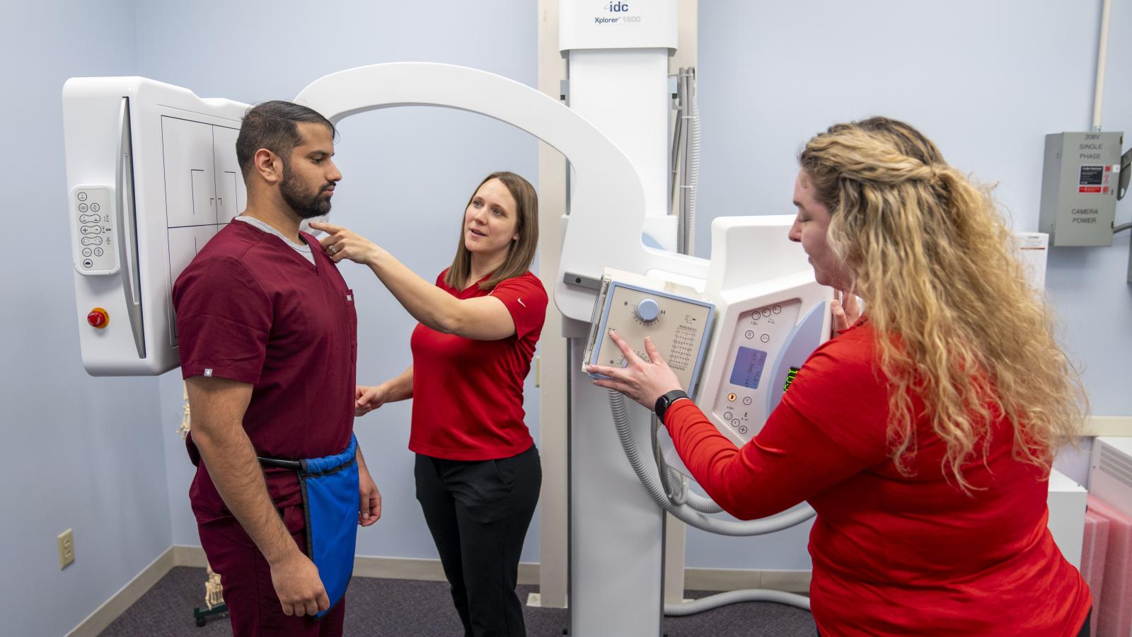 Three people in a room with x-ray machine. Man stands for an x-ray. one woman is operating the machine while the other is pointing and teaching.