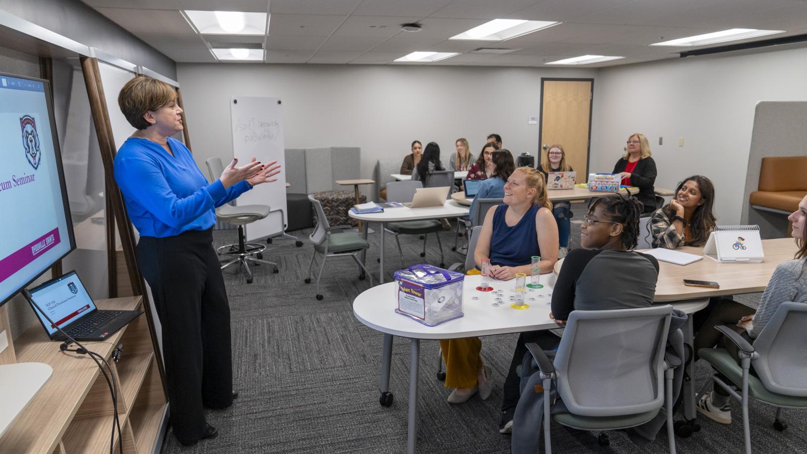 woman stands at the front of a classroom speaking to a large group.