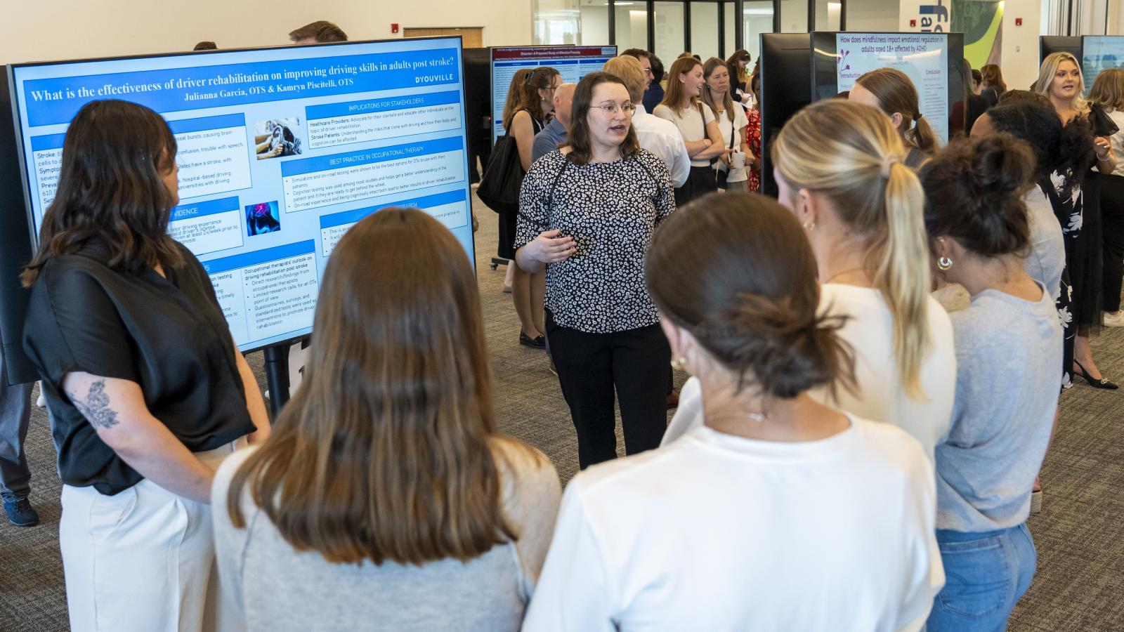 Student stands next to a large television display their research. a group listens while student speaks.