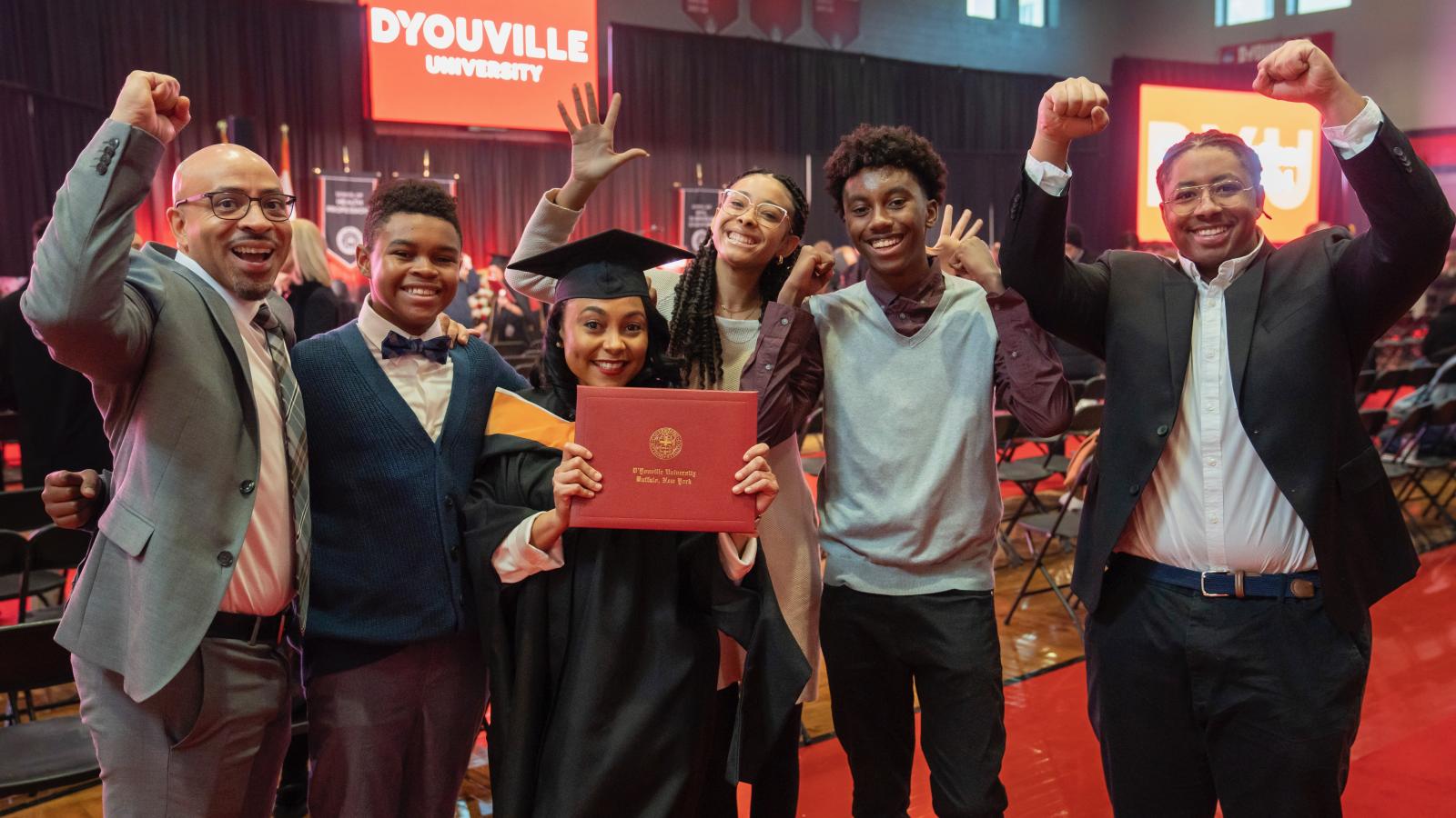 Student in cap and gown holding a diploma stands at the center of a row of people cheering and raising their arms in celebration