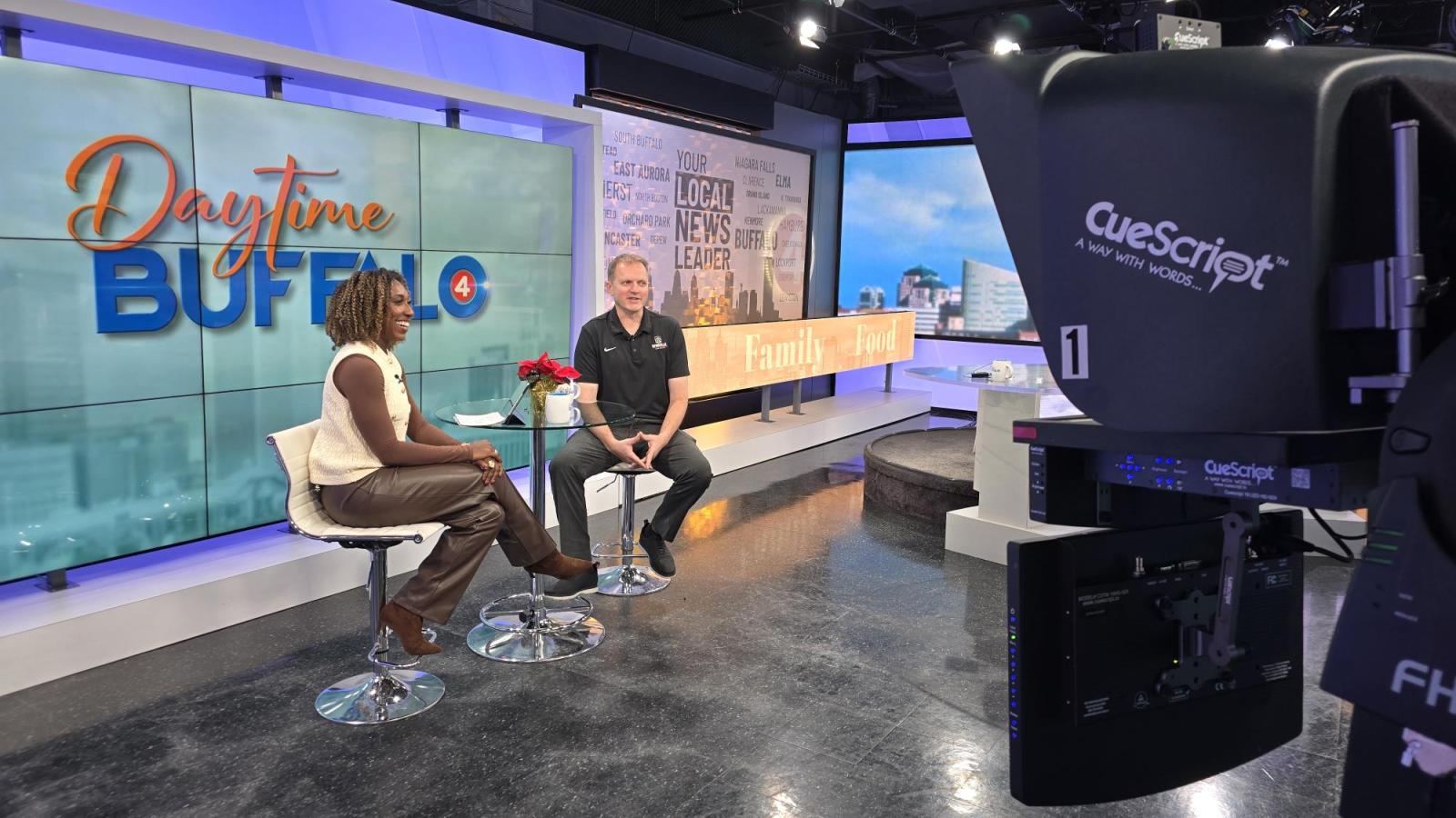 two people sit at a table and chairs inside a television studio. a camera is in the foreground of the image.