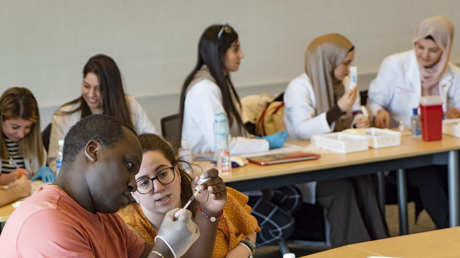 in the foreground you see two people, one holds a needle and is drawing liquid from a bottle. in the background you see five students sitting at a table.
