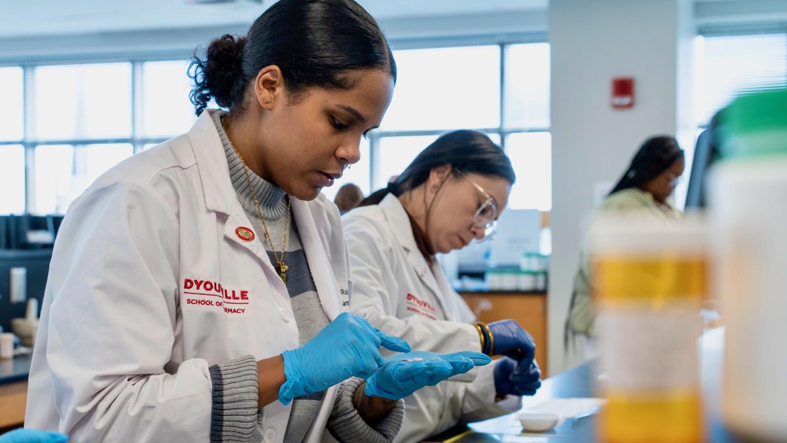 Two women standing at a table in white coats. The patch on the white coat reads D'Youville School of Pharmacy. The women in the foreground is counting a handful of pills.