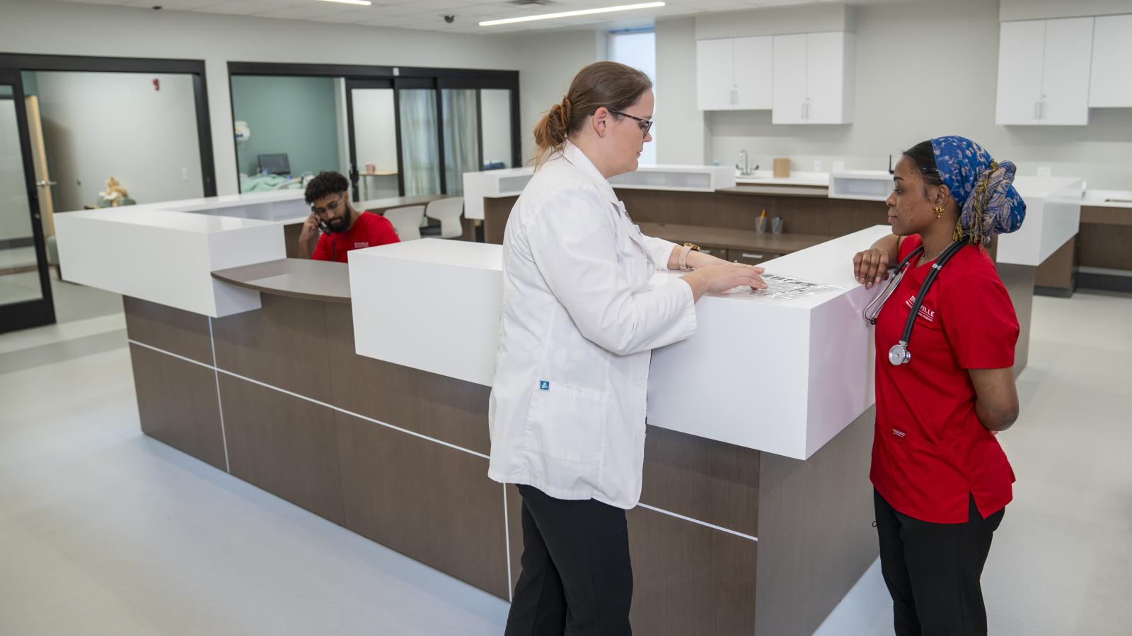 two people stand in front of a nurses station reviewing a document.