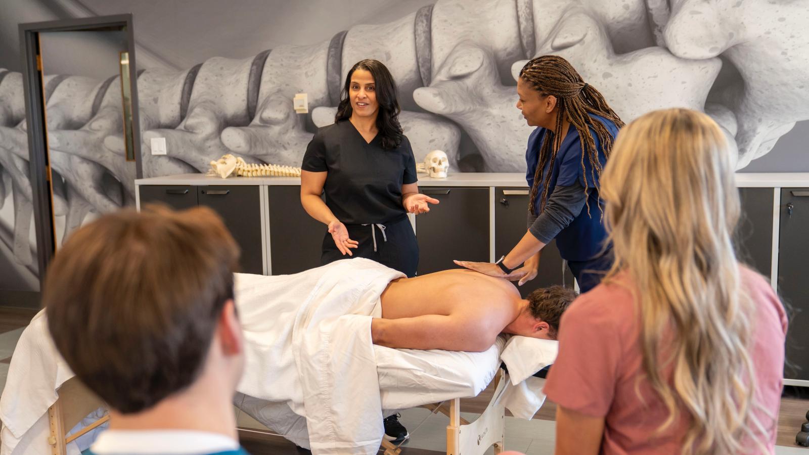 one student lies on a massage table being worked on by another student while an instructor speaks to the class
