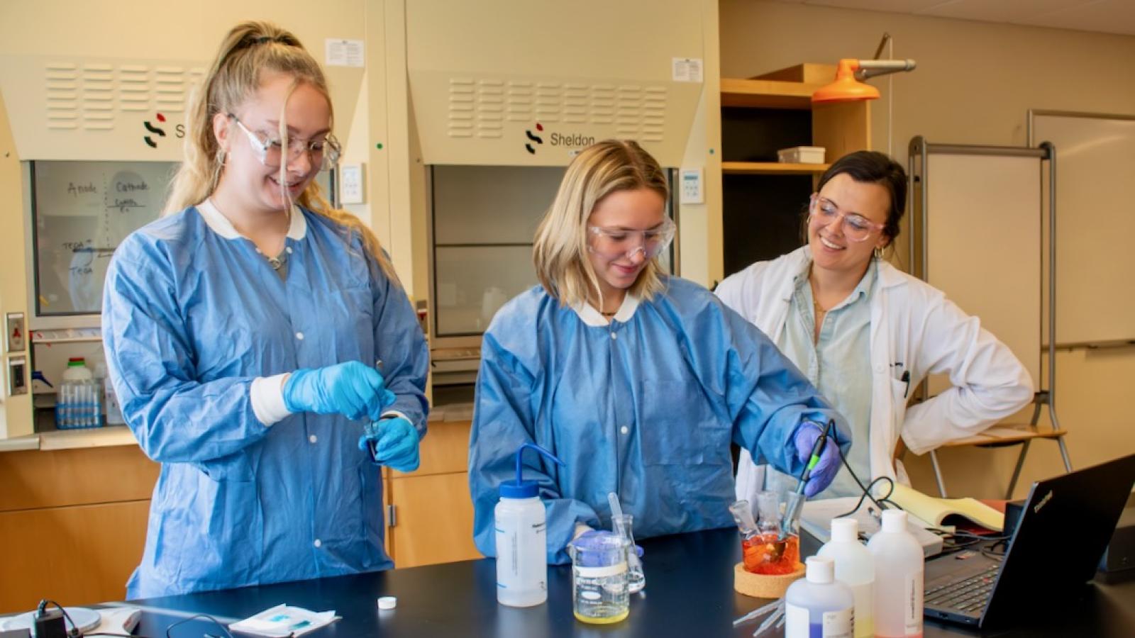 Three women stand in a lab at a table. One woman is using a pipette to move liquid into a beaker.