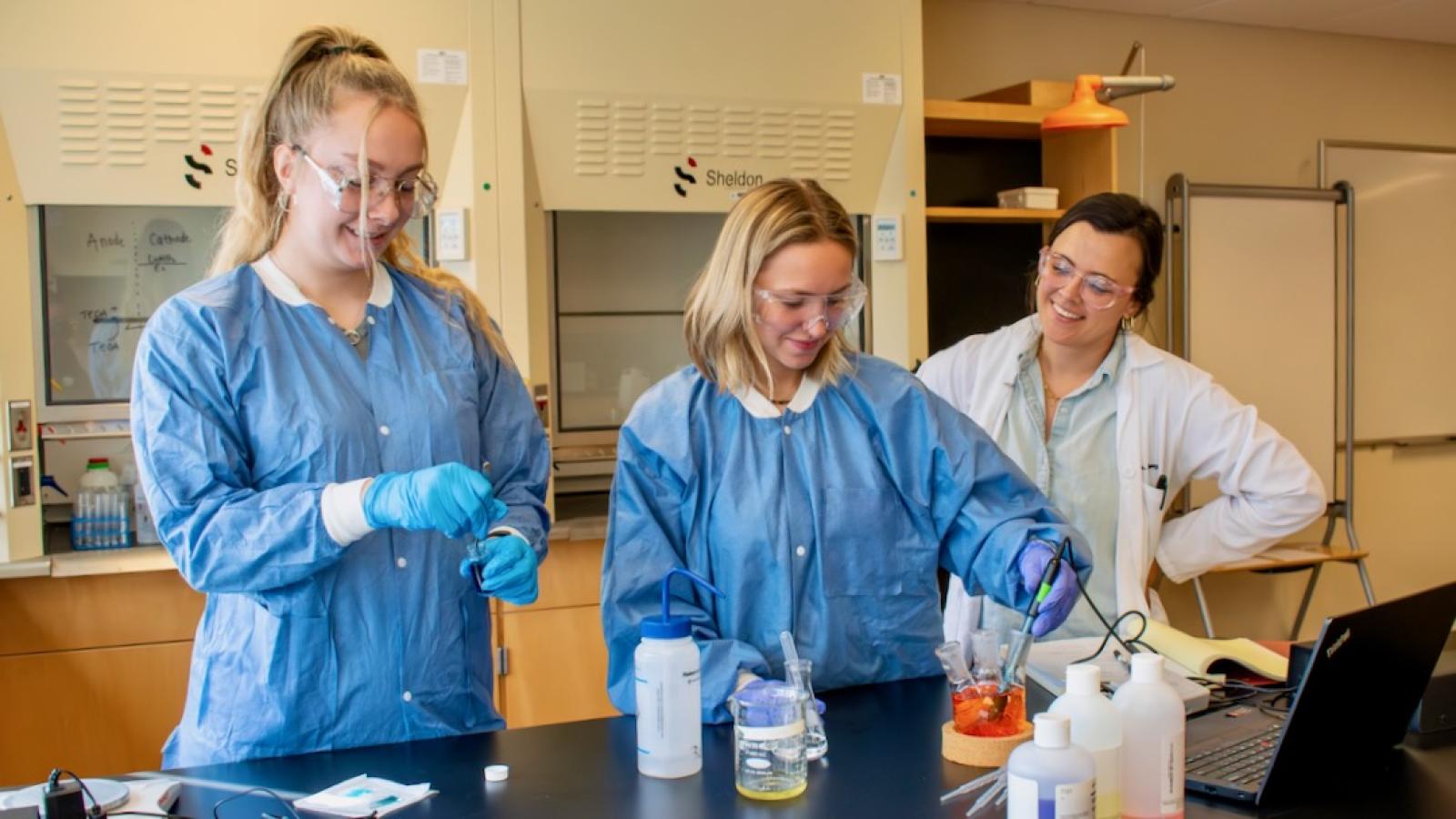 Three women stand in a lab at a table. One woman is using a pipette to move liquid into a beaker.