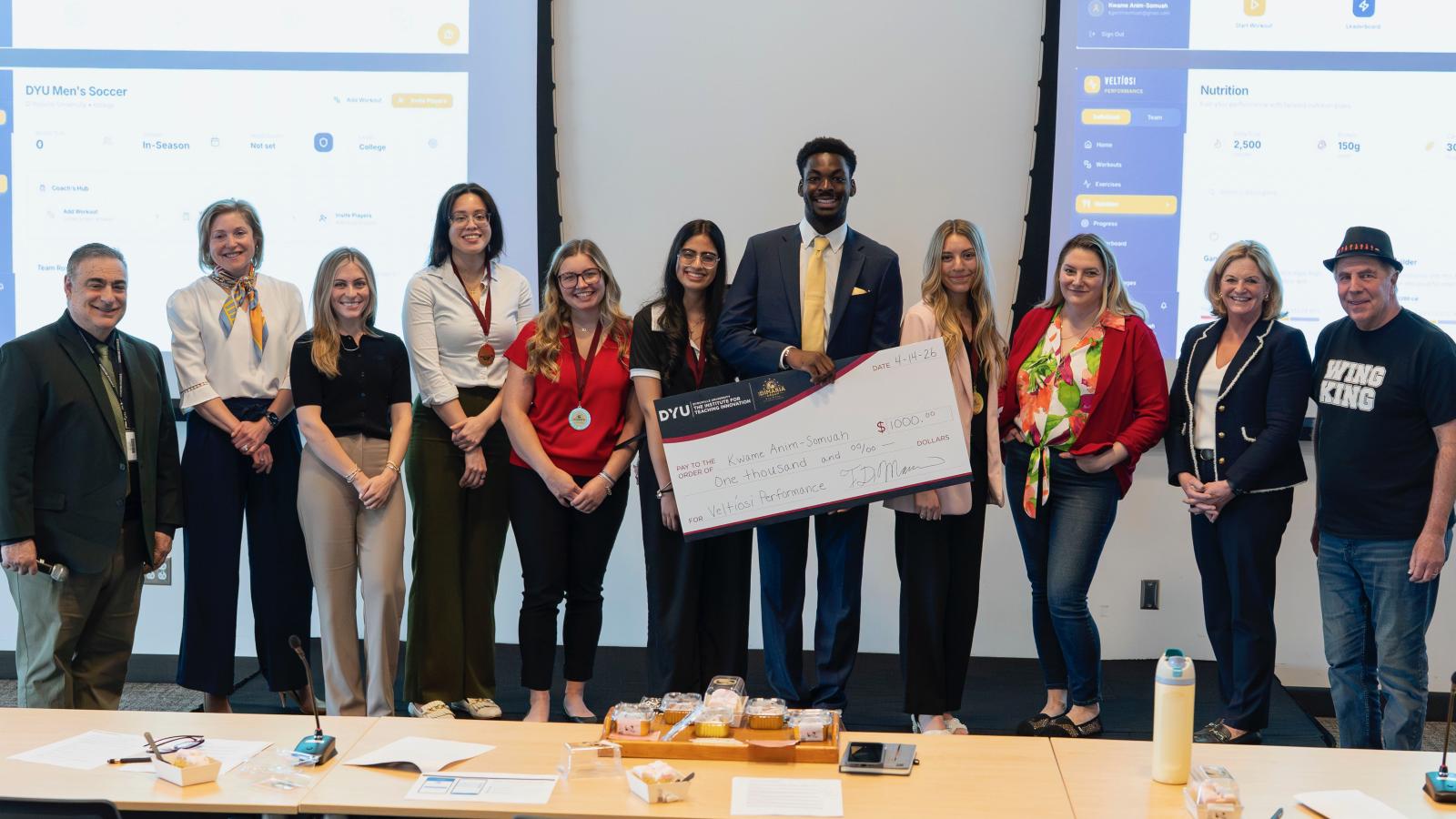 group stands in a row smiling at the camera. One stands holding an oversized check.