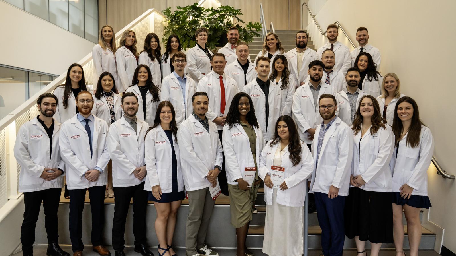 large group of people stand on steps smiling at the camera wearing white coats