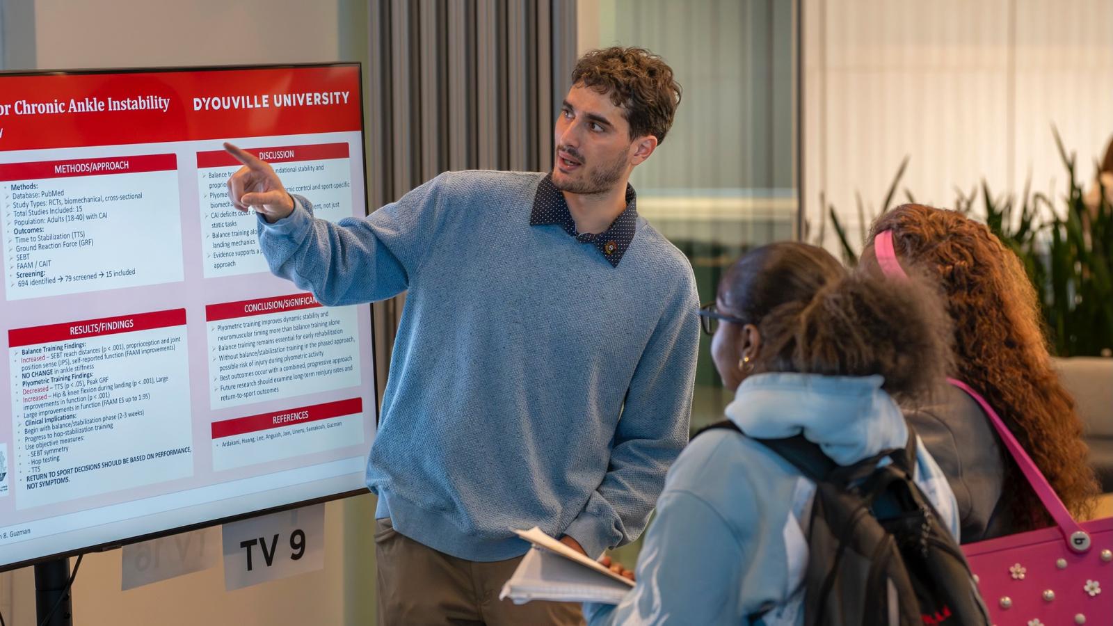 student points at a research poster while speaking to two fellow students