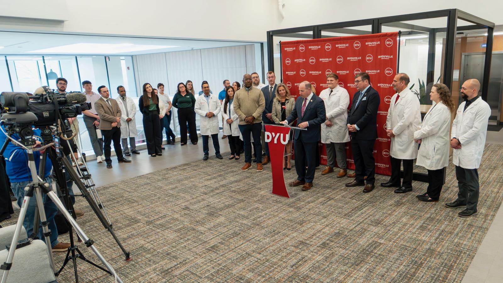 large group of people stand in an atrium. tim kennedy stands at a podium addressing the crowd.