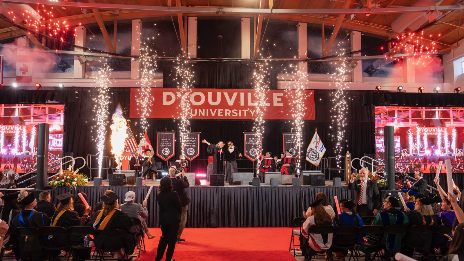 large indoor stage with sparks and fireworks. You see a large banner in the background that reads D'Youville University.