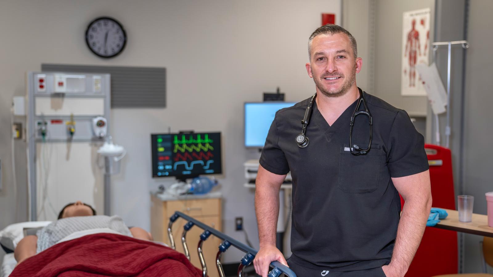 man in scrubs stands smiling in front of a medical simulation room set up like an ER