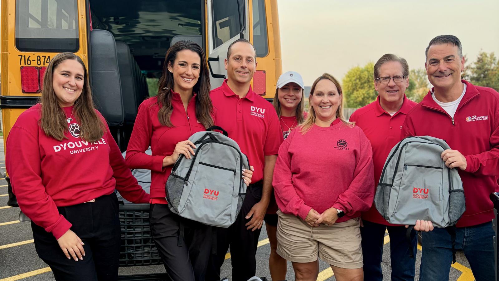 group of people stand smiling at the camera. Two people are holding up backpacks that read: DYU D'Youville University