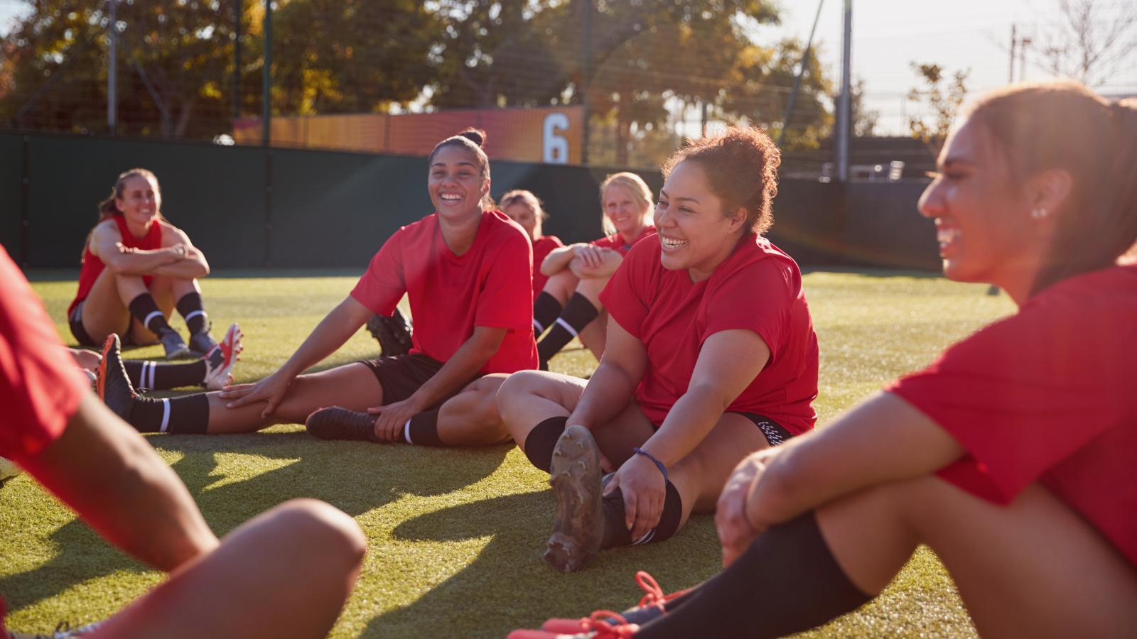group of women in soccer uniforms stretching on a field, laughing and smiling