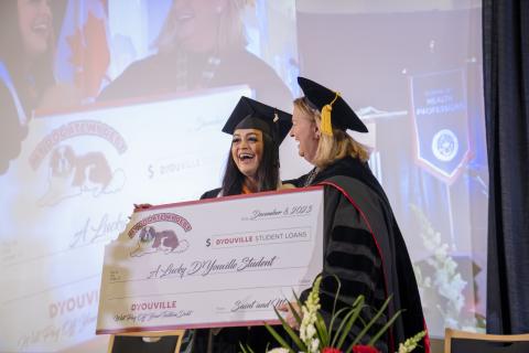President Dr. Lorrie Clemo, in graduation regalia, stands with student in graduation cap and gown. They hold a giant check representing a payment of the lottery winner's student loan debt.