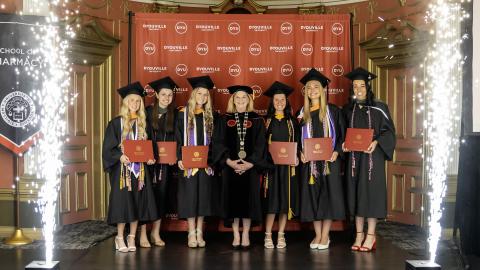 six 2025 graduates from the dyouville university softball team stand with president lorrie clemo with pyrotechnic sparks on either side.