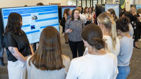 Student stands next to a large television display their research. a group listens while student speaks.