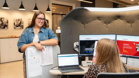 one woman stands smiling down at another who is not facing the camera sitting at her desk