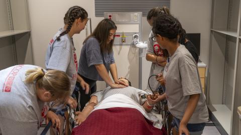 five students stand around a hospital bed. in the bed is a manikin and one student is practicing chest compressions.