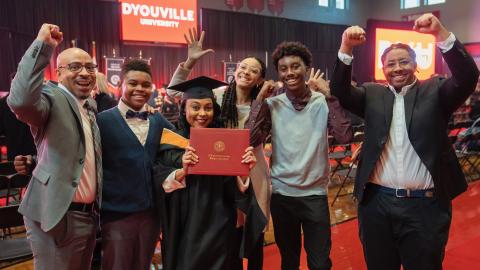 Student in cap and gown holding a diploma stands at the center of a row of people cheering and raising their arms in celebration