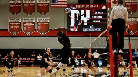 womens volleyball game viewed from mid-court. One woman jumps to spike a ball.