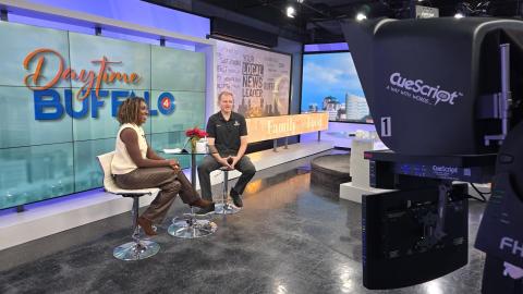two people sit at a table and chairs inside a television studio. a camera is in the foreground of the image.