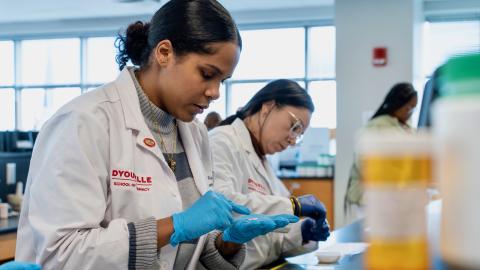 Two women standing at a table in white coats. The patch on the white coat reads D'Youville School of Pharmacy. The women in the foreground is counting a handful of pills.