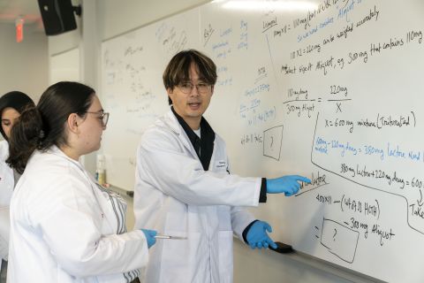 two students in white coats stand talking in front of a large white board with mathematic equations on it. 