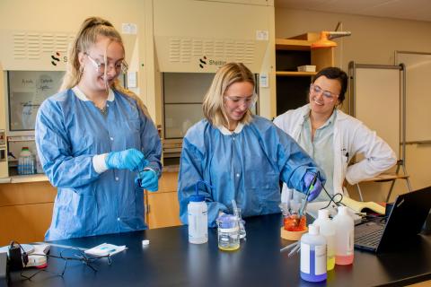 Three women stand at a lab table in a chemistry lab. There are several bottles and beakers of colored liquid on the table. 