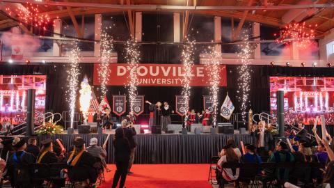 large indoor stage with sparks and fireworks. You see a large banner in the background that reads D'Youville University.