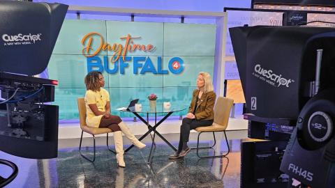 Two women sit at a table on a TV set. You see television station cameras in the foreground.