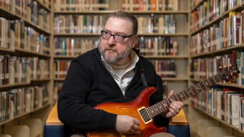 man sitting in chair playing a guitar, bookcases in the background