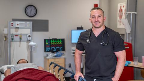 man in scrubs stands smiling in front of a medical simulation room set up like an ER