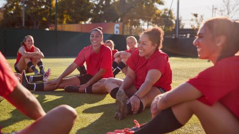 group of women in soccer uniforms stretching on a field, laughing and smiling