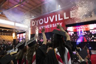 Students shown from behind in graduation caps cheering. fireworks are happening on a stage with a large D'Youville University banner above it..