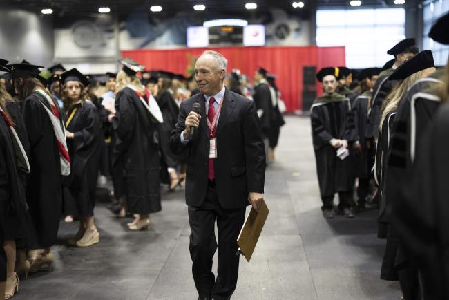 Man walking with microphone in a room with students in graduation regalia