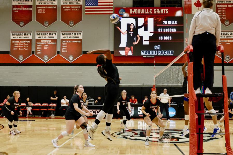 inside a gym looking at the court of a women's volleyball match. One player is jumping in the air to spike the ball.