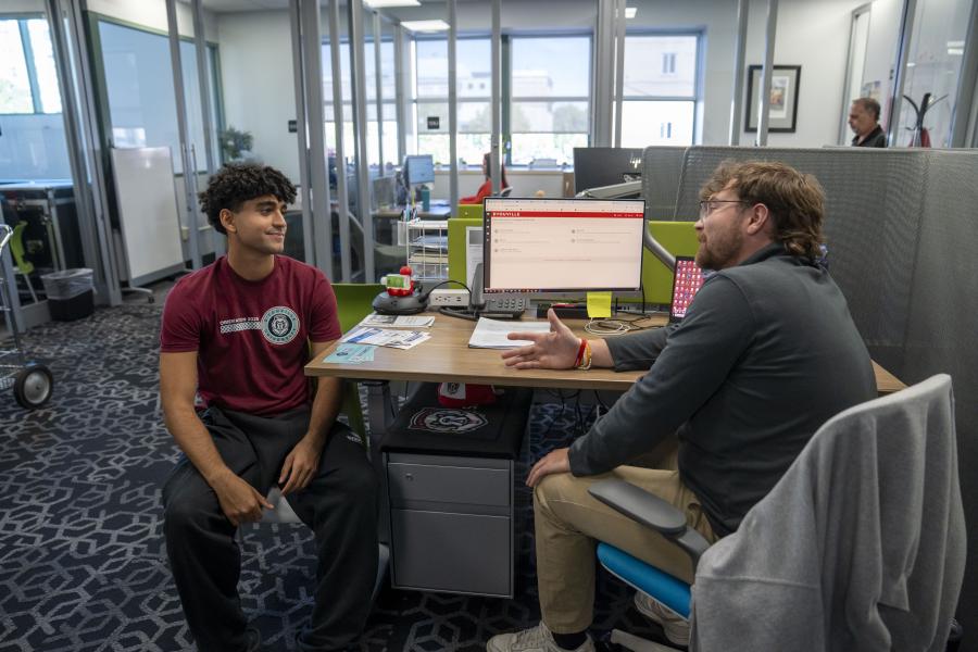 two men sit in a large office talking across a desk from one another.