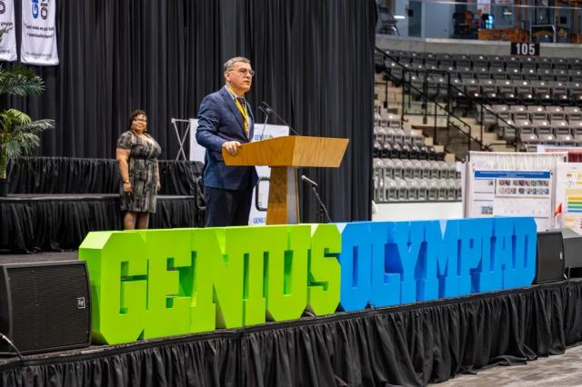 Dr. Fehmi Damkaci stands at podium on a stage behind a Genius Olympiad sign