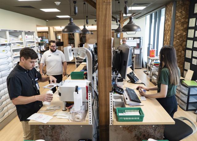 Two young men and a woman working inside a pharmacy. One fills a pill bottle, another works on a computer, and the third is walking in the room.