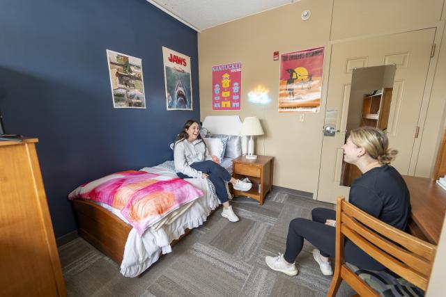 Two women in residence hall room. One is sitting on bed and one on a desk chair.