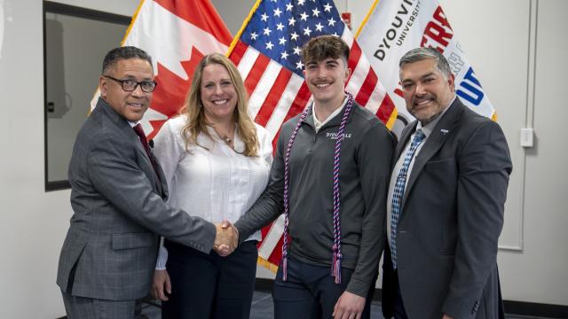 four people stand and smile at the camera. student wears veteran graduation cord and shakes hands with the veterans office director