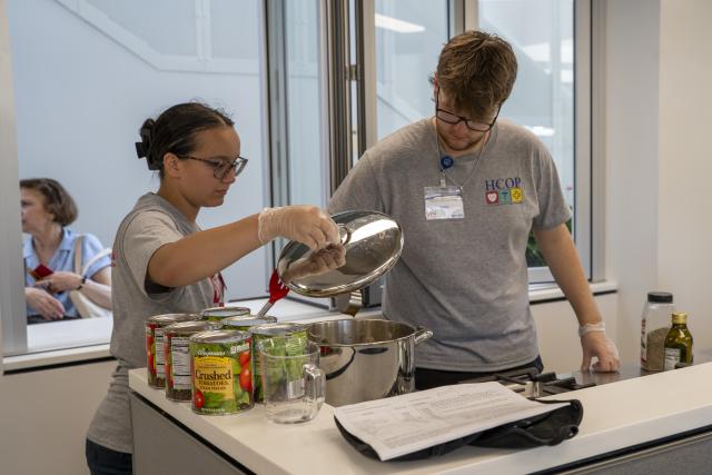two young people stand at a cooktop. one is lifting the lid off of a pot.