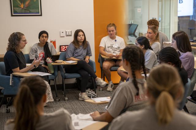a large group of students sit in a circle in a classroom. a professor is speaking.