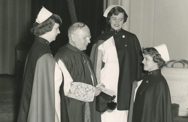 Priest stands with three nursing students at a graduation ceremony