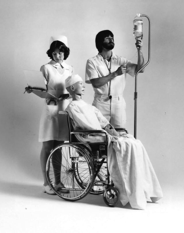 man in scrubs stands a iv pole. woman nurse stands with clipboard. both are behind a maniquin in a wheelchair.